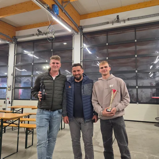 Drei Männer stehen in einer Einrichtung, einer hält eine Trophäe und der andere eine Flasche. Der Mann auf der linken Seite lächelt, während die anderen für ein Foto posieren. Hinter ihnen sind Tische und große Fenster.