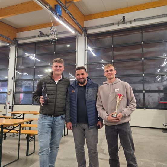 Drei Männer stehen in einer Einrichtung, einer hält eine Trophäe und der andere eine Flasche. Der Mann auf der linken Seite lächelt, während die anderen für ein Foto posieren. Hinter ihnen sind Tische und große Fenster.