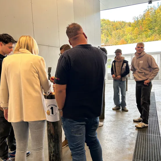 Eine Gruppe von Menschen steht drinnen versammelt, wobei eine Person einen Kürbis mit der Nummer 1 schnitzt. Sie stehen vor einer Betonwand mit einem Glasfenster, das Herbstbäume zeigt.