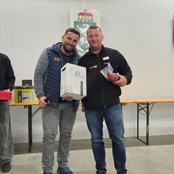 Zwei Männer stehen in einem Raum zusammen, einer hält ein Mikrofon und der andere eine Box mit Paschek-Wein. Eine dritte Person steht hinter ihnen. Der Raum hat Tische, Stühle und eine Wand mit einem Wappen.
