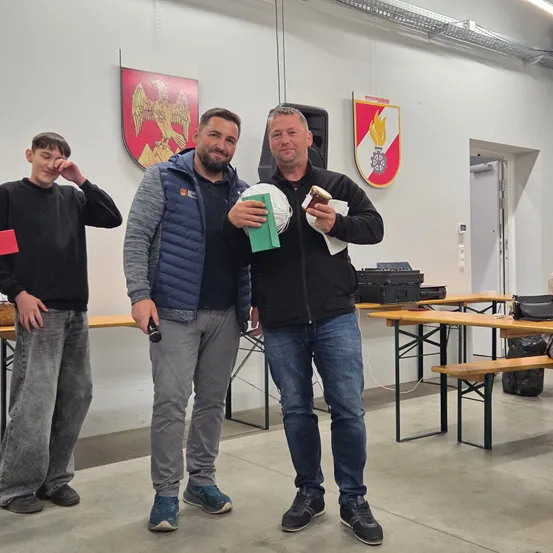 Drei Männer stehen in einem Raum, einer hält eine grüne Mappe und einen Fußball, ein anderer ein Mikrofon, und ein Junge hält eine rote Karte. Sie stehen vor Tischen und Stühlen, mit Wappen an der Wand.