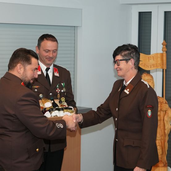 Drei Menschen in Uniform stehen. Der Mann in der Mitte hält eine Schachtel mit Flaschen. Die anderen beiden schütteln Hände. Dahinter steht eine Holzstruktur mit einer Fahne.