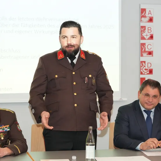 Ein Mann in Uniform steht vor einer Leinwand, möglicherweise gibt er eine Präsentation. Zwei andere Männer, einer in Anzug, sitzen hinter einem Tisch mit einer Flasche und Papieren.