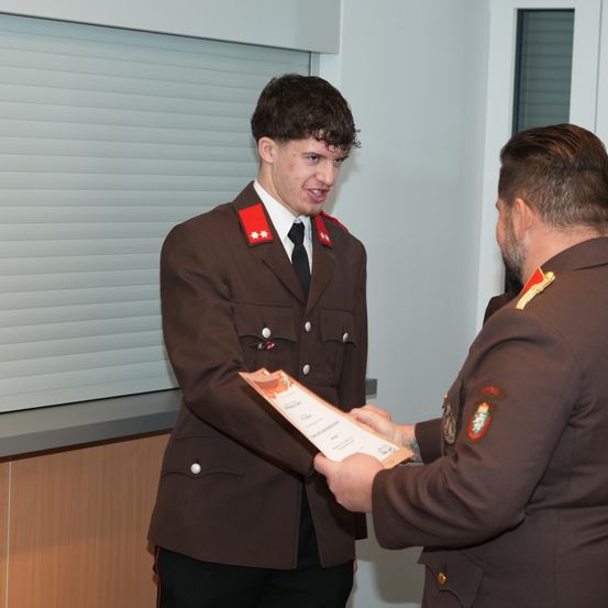 Ein Mann in Uniform überreicht einem anderen Mann in ähnlicher Uniform ein Zertifikat. Sie stehen drinnen.