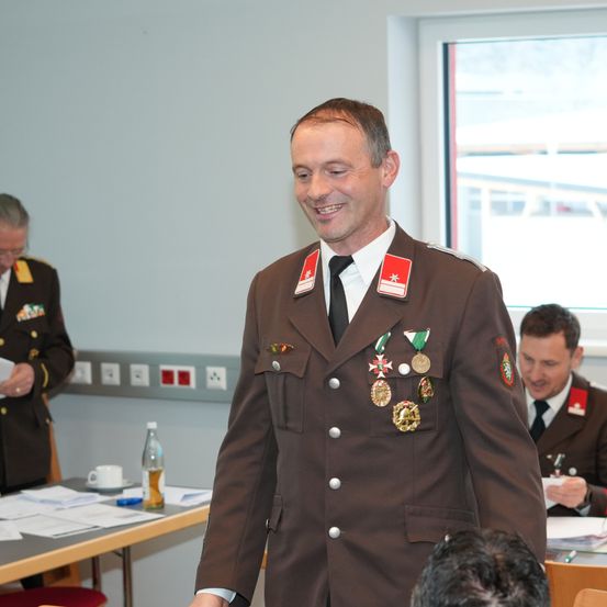Ein Mann in Uniform mit Medaillen lächelt in einem Raum mit anderen Militäroffizieren, einem Schreibtisch und einem Fenster.