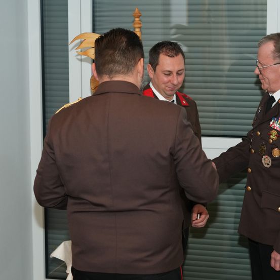 Drei Männer in Militäruniformen stehen in einem Raum, zwei scheinen zu reden, während der Dritte zusieht. Der Raum hat eine weiße Wand und ein Fenster mit Jalousien.