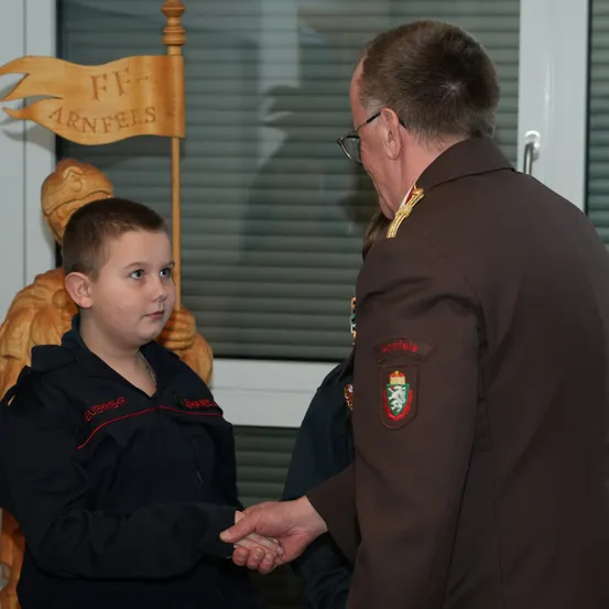 Ein uniformierter Mann schüttelt die Hand eines jungen Jungen, der einen schwarzen Mantel mit rotem Saum trägt. Hinter ihnen befindet sich ein Holzbanner mit dem Text 'FF Arnfels'.