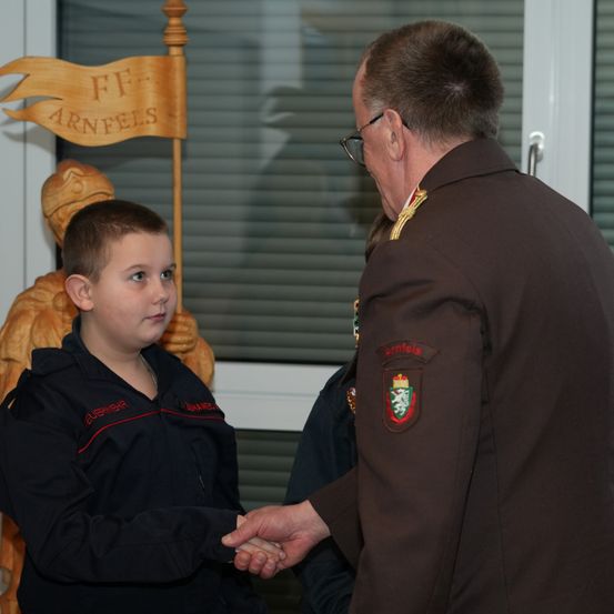 Ein uniformierter Mann schüttelt die Hand eines jungen Jungen, der einen schwarzen Mantel mit rotem Saum trägt. Hinter ihnen befindet sich ein Holzbanner mit dem Text 'FF Arnfels'.