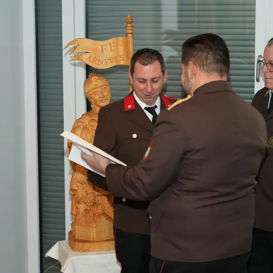 Drei Männer in Militäruniformen stehen in einem Raum. Einer hält ein Stück Papier und schaut darauf, während die anderen beiden ihn ansehen. Hinter ihnen steht eine Holzstatue mit einer Flagge oben.
