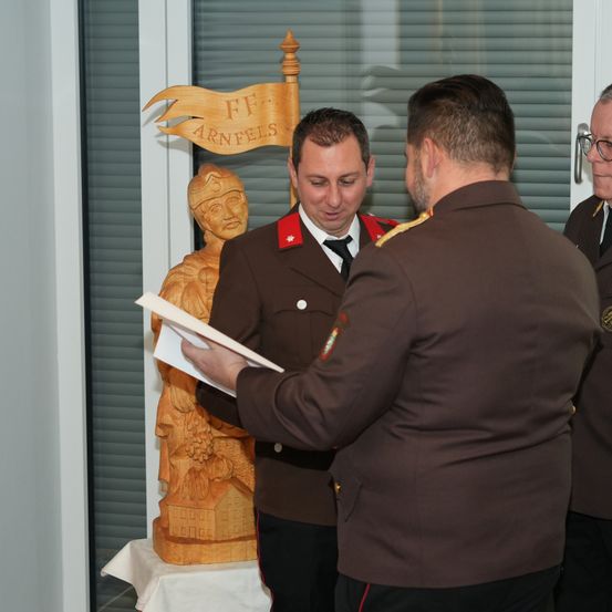 Drei Männer in Militäruniformen stehen in einem Raum. Einer hält ein Stück Papier und schaut darauf, während die anderen beiden ihn ansehen. Hinter ihnen steht eine Holzstatue mit einer Flagge oben.