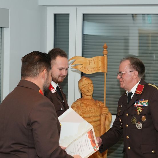 Drei Männer in Uniformen stehen vor einer Statue, einer hält ein Papier, ein anderer schüttelt die Hand eines Mannes in einer dekorierten Militäruniform. Dahinter ist ein Holzschild mit den Buchstaben FF Arnfels zu sehen.