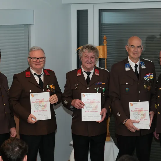 Vier Männer in Uniformen stehen zusammen, lächeln und halten Urkunden in den Händen. Hinter ihnen schaut ein weiterer Mann zu. Sie befinden sich in einem Raum mit weißen Wänden und Fenstern mit Jalousien.