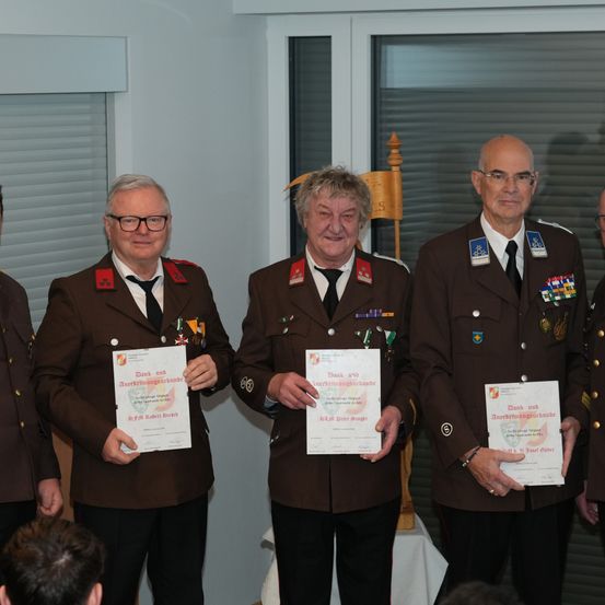 Vier Männer in Uniformen stehen zusammen, lächeln und halten Urkunden in den Händen. Hinter ihnen schaut ein weiterer Mann zu. Sie befinden sich in einem Raum mit weißen Wänden und Fenstern mit Jalousien.