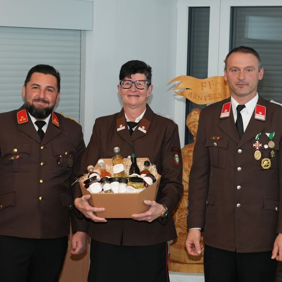 Drei Menschen in Militäruniformen stehen zusammen und lächeln. Die Frau in der Mitte hält eine Box mit verschiedenen Flaschen. Dahinter steht eine Holzstatue und ein Schild mit dem Text 'F.F. Arnfels'.