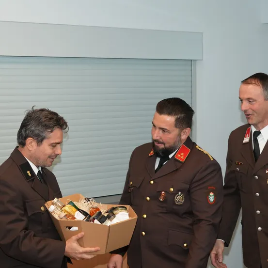 Drei Männer in Militäruniformen stehen. Der Mann auf der linken Seite hält eine Kiste voller verschiedener Gegenstände. Der Mann in der Mitte schaut auf die Kiste. Der Mann auf der rechten Seite lächelt. Hinter ihnen befindet sich eine weiße Wand mit einem Fenster.