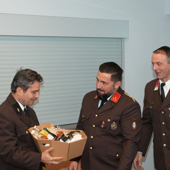 Drei Männer in Militäruniformen stehen. Der Mann auf der linken Seite hält eine Kiste voller verschiedener Gegenstände. Der Mann in der Mitte schaut auf die Kiste. Der Mann auf der rechten Seite lächelt. Hinter ihnen befindet sich eine weiße Wand mit einem Fenster.