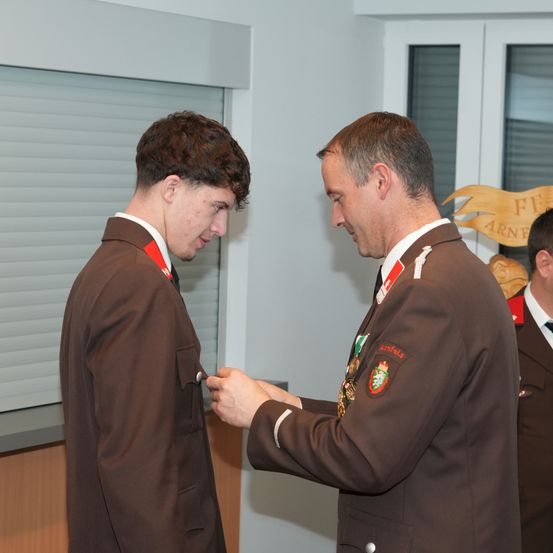 Zwei Männer in braunen Uniformen sind drinnen, einer befestigt eine Medaille am anderen. Hinter ihnen zeigt eine Wand ein Holzschild mit Text und einem Emblem.