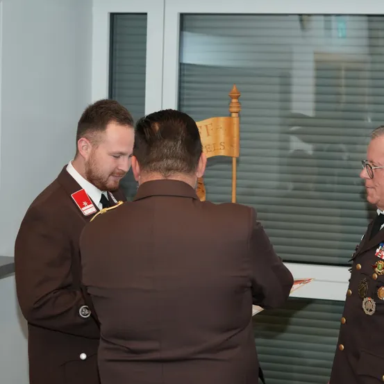 Drei Männer in Militäruniformen stehen in einem Innenraum. Einer liest ein Papier, ein anderer schaut ihn an, und der dritte steht hinter einem Holzschild. Ein Glasfenster befindet sich hinter ihnen.
