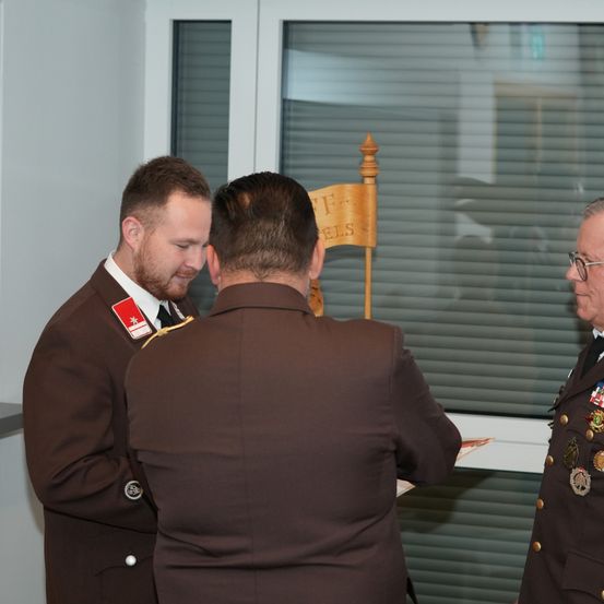 Drei Männer in Militäruniformen stehen in einem Innenraum. Einer liest ein Papier, ein anderer schaut ihn an, und der dritte steht hinter einem Holzschild. Ein Glasfenster befindet sich hinter ihnen.