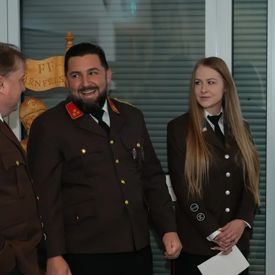 Drei Personen in Militäruniformen stehen vor einem Gebäude mit einem Banner, das FF-KARNFEL steht. Ein Mann lächelt, eine Frau hält ein Stück Papier und ein anderer Mann schaut zur Seite.