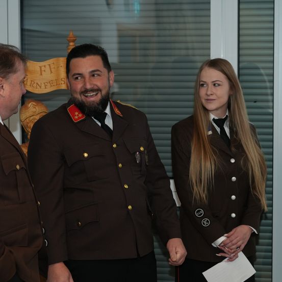 Drei Personen in Militäruniformen stehen vor einem Gebäude mit einem Banner, das FF-KARNFEL steht. Ein Mann lächelt, eine Frau hält ein Stück Papier und ein anderer Mann schaut zur Seite.