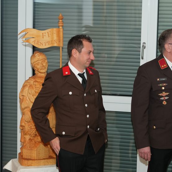 Zwei Männer in Militäruniformen stehen drinnen in der Nähe eines Fensters. Einer hat die Hände in den Taschen und lächelt. Hinter ihm hält eine Holzstatue eine Fahne.