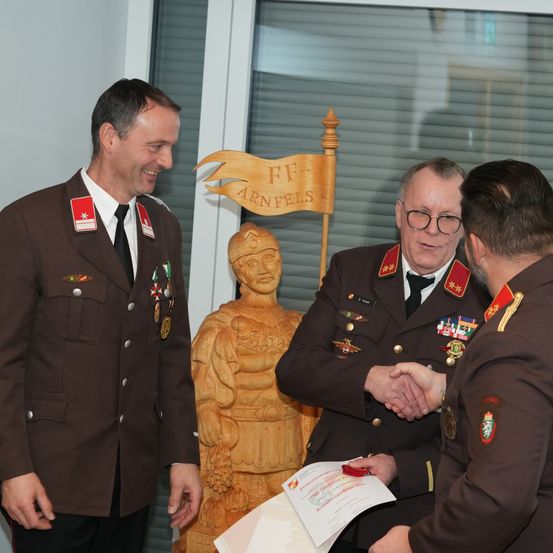 Drei Männer in Militäruniformen tauschen einen Händedruck aus. Hinter ihnen steht eine Holzstatue, und einer der Männer hält ein Zertifikat. Eine Fahne mit FF Arnfels ist zu sehen.