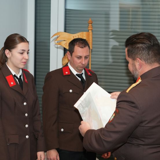 Drei Menschen in Militäruniformen stehen neben einem hölzernen Objekt. Ein Mann in der Mitte hält ein Stück Papier. Eine Frau schaut den Mann auf der linken Seite an.