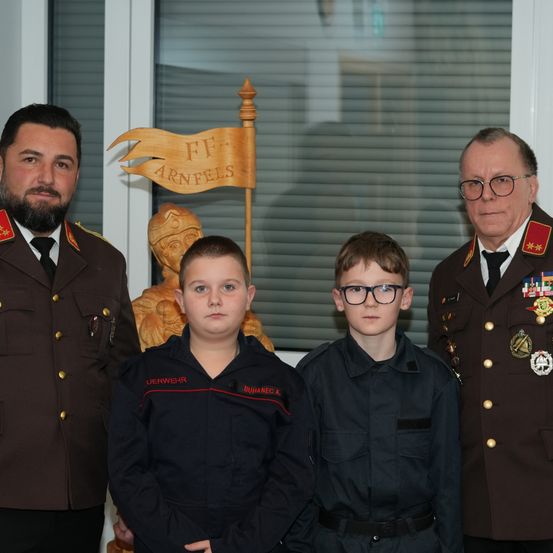 Zwei uniformierte Jungen stehen neben zwei Männern in Militäruniform, einer mit Brille, alle vor einem hölzernen Banner.