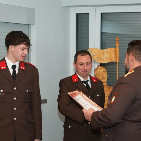 Drei Männer in Militäruniformen stehen in einem Raum. Einer hält ein Zertifikat, ein anderer schaut darauf. Eine Statue und ein Schild mit 'FF ARNFELS' sind dahinter.