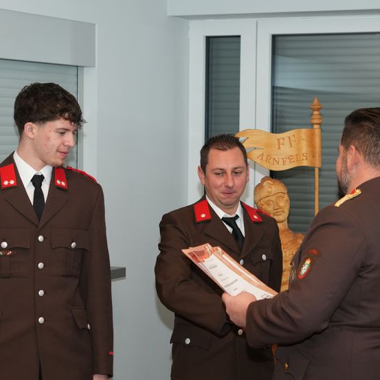 Drei Männer in Militäruniformen stehen in einem Raum. Einer hält ein Zertifikat, ein anderer schaut darauf. Eine Statue und ein Schild mit 'FF ARNFELS' sind dahinter.