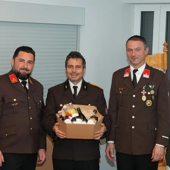 Drei Männer in Militäruniformen stehen nebeneinander. Der Mann in der Mitte hält eine Box mit Flaschen. Sie stehen in einem Raum mit einer weißen Wand und einem Holzstuhl.