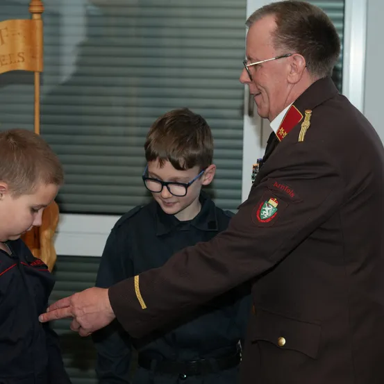 Ein uniformierter Mann mit Brille überreicht zwei jungen Jungen in einem Raum eine Auszeichnung.