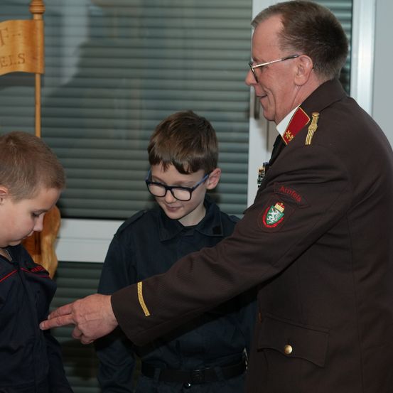 Ein uniformierter Mann mit Brille überreicht zwei jungen Jungen in einem Raum eine Auszeichnung.