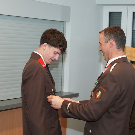 Ein Mann in Uniform befestigt eine Anstecknadel am Jackett eines jüngeren Mannes, beide lächeln einander an.