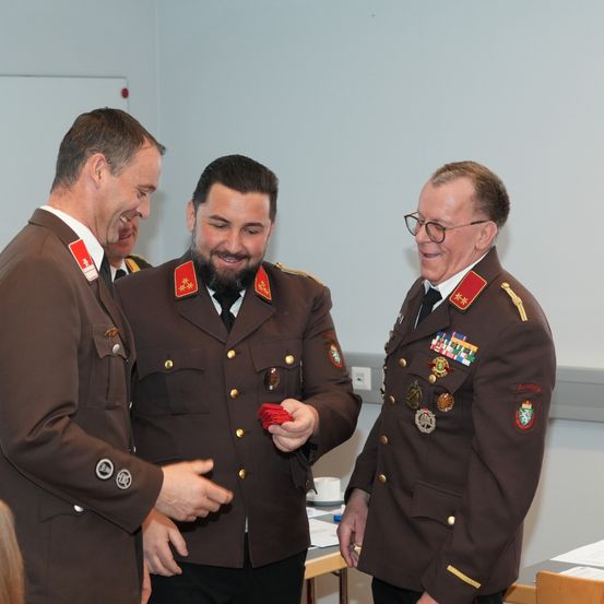 Drei Männer in Militäruniformen stehen in einem Raum, einer hält rote Karten. Sie lächeln und scheinen zu sprechen.