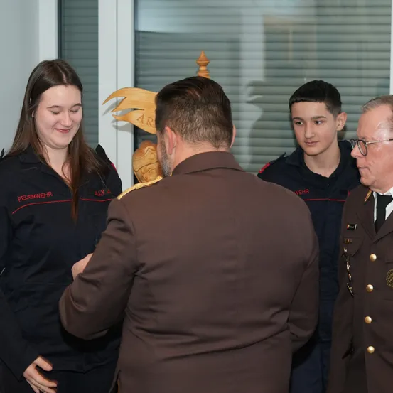 Drei uniformierte Personen stehen vor einem Mann in einem braunen Anzug. Die Frau und der Junge lächeln, während der ältere Mann eine Brille trägt. Ein Holzpfahl steht hinter ihnen.