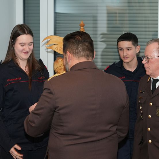 Drei uniformierte Personen stehen vor einem Mann in einem braunen Anzug. Die Frau und der Junge lächeln, während der ältere Mann eine Brille trägt. Ein Holzpfahl steht hinter ihnen.