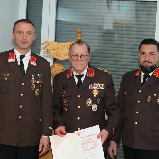 Drei Männer in Militäruniformen stehen nebeneinander, wobei der mittlere Mann ein Zertifikat in der Hand hält. Sie stehen vor einer Wand mit einem Holzschild und Jalousien.