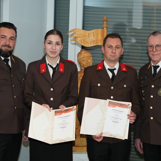 Vier Personen in Militäruniformen, zwei Männer und zwei Frauen, stehen zusammen und halten Urkunden. Sie stehen vor einem hölzernen Banner.