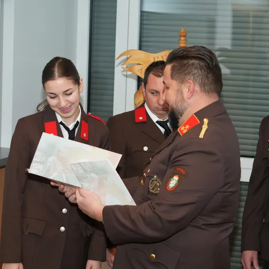 Eine lächelnde Frau in einer braunen Uniform hält ein Dokument, während ein Mann in Uniform es betrachtet. Hinter ihnen schaut ein weiterer uniformierter Mann zu, und ein hölzerne Emblem hängt über ihnen.