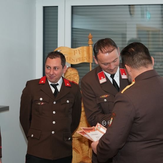 Drei Männer in Militäruniformen stehen neben einem Holzstuhl, einer hält ein Zertifikat, ein anderer liest es, und der dritte schaut zu. Der Stuhl trägt die Worte 'FF Warnfels'.