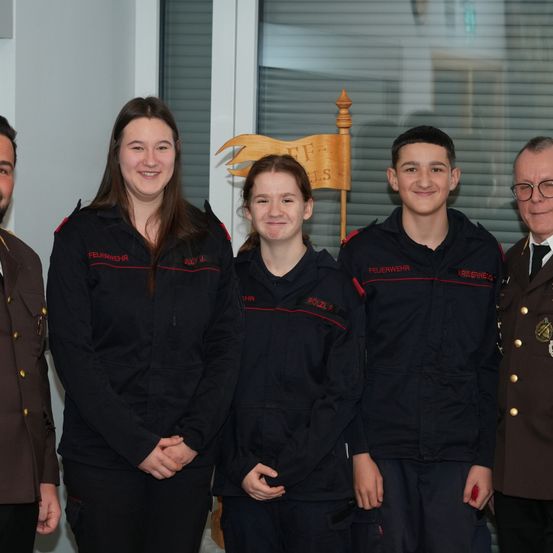 Fünf Personen stehen in einer Reihe und lächeln für ein Foto. Die Frau auf der linken Seite trägt eine Jacke mit dem Wort Feuerwehr. Dahinter steht ein Holzschild mit dem Text TFLS. Der Mann auf der rechten Seite trägt eine Brille und eine Militäruniform.