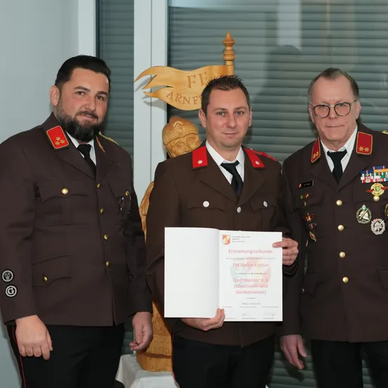 Drei Männer in Militäruniformen stehen eng beieinander. Der Mann in der Mitte hält ein Zertifikat. Hinter ihnen ist ein Holzbanner mit den Buchstaben FF ArnF.