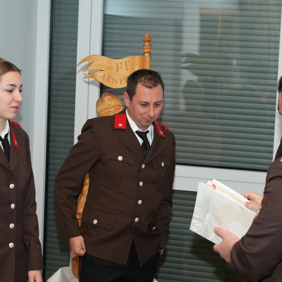 Ein Mann in einer braunen Uniform steht zwischen zwei anderen und schaut auf ein Dokument. Hinter ihm steht ein hölzernes Banner mit der Aufschrift FF. Eine Frau in einer ähnlichen Uniform steht zu seiner Linken.
