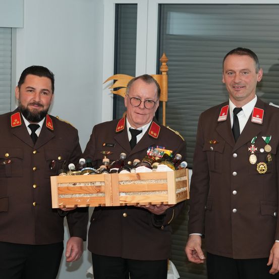 Drei Männer in Militäruniformen, einer hält eine Holzkiste voller Flaschen, stehen nebeneinander in einem Raum.