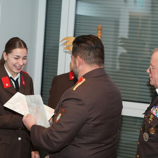 Eine Frau in Uniform hält eine Karte und lächelt, während ein Mann in Uniform ihr ein Dokument reicht. Ein weiterer Mann in Uniform steht hinter ihnen.