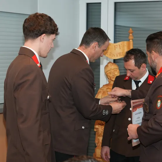 Vier Männer in Militäruniformen stehen in geschlossenen Räumen, einer befestigt eine Medaille an einem anderen. Der Mann, der die Medaille erhält, sieht nach unten, und der Mann, der die Medaille befestigt, hält ein Papier. Eine Holzstatue ist im Hintergrund.