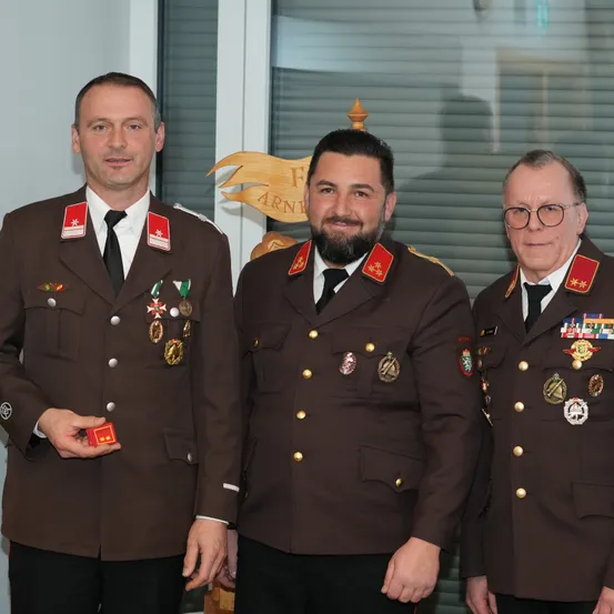 Drei Männer in Militäruniformen, eng beieinander stehend, lächeln für die Kamera. Der Mann auf der linken Seite hält eine kleine rote Schachtel. Hinter ihnen befindet sich eine Wand mit einem goldenen Design und einem Schild.