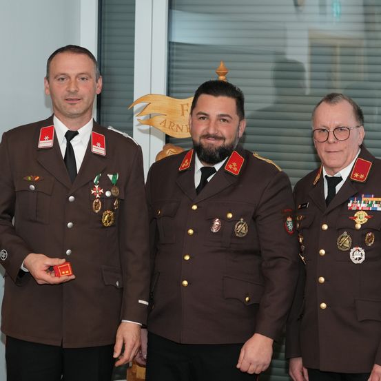 Drei Männer in Militäruniformen, eng beieinander stehend, lächeln für die Kamera. Der Mann auf der linken Seite hält eine kleine rote Schachtel. Hinter ihnen befindet sich eine Wand mit einem goldenen Design und einem Schild.